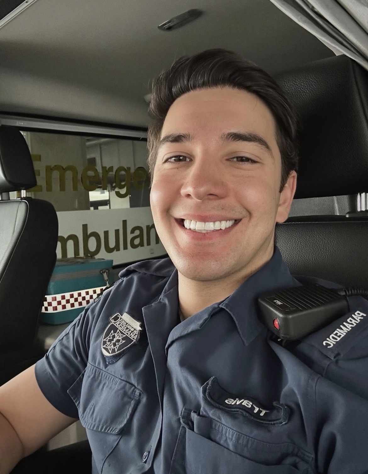Close-up of Travis Wishart smiling in the front seat of an ambulance, wearing a dark blue NSW Ambulance paramedic uniform with a radio microphone on his shoulder. The word “Emergency Ambulance” is partially visible on the glass behind him, along with medical equipment in the background.