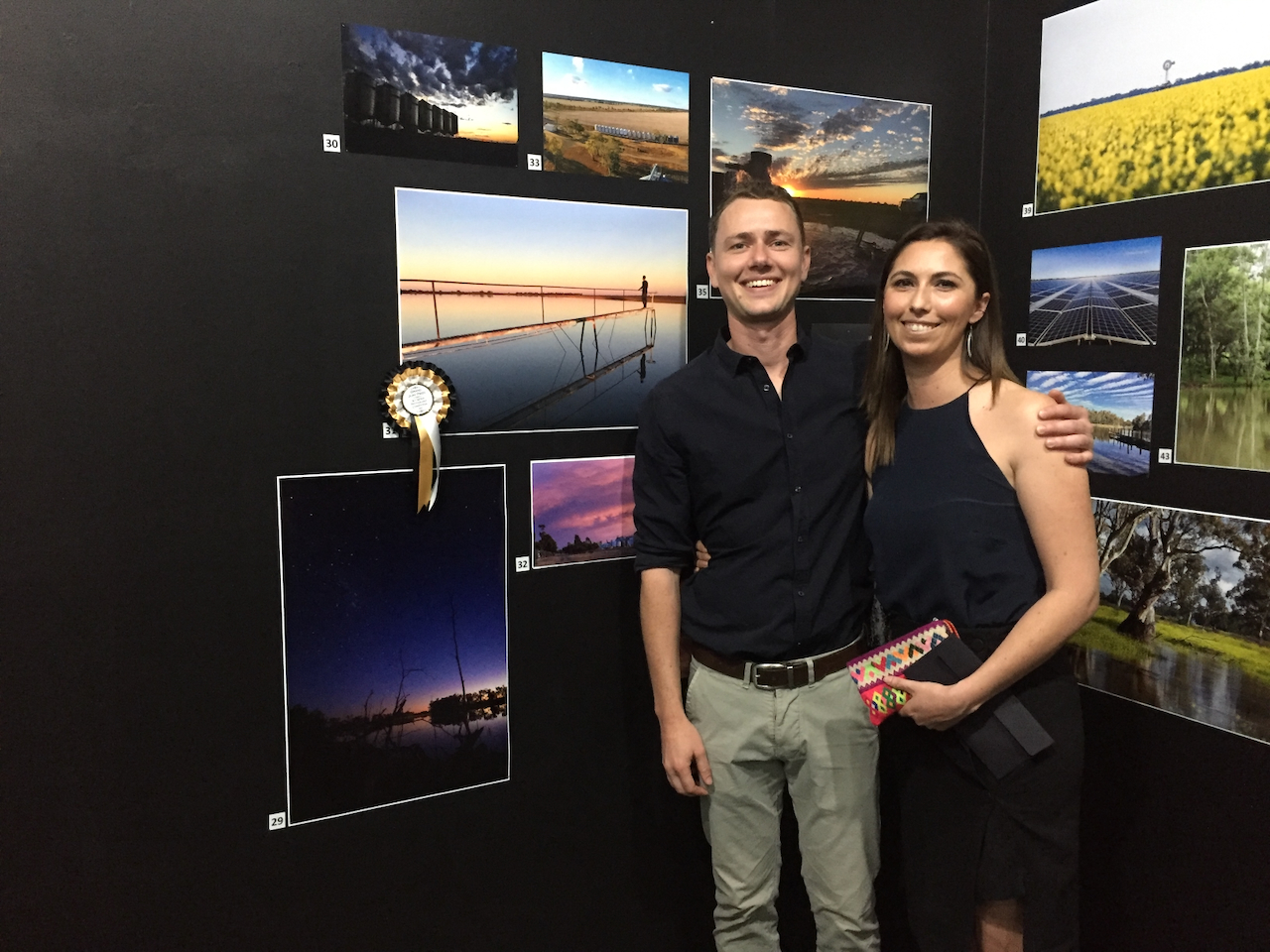 Two adults stand smiling in front of a black exhibition wall covered with framed landscape photographs; an award ribbon is pinned beside one of the displayed images.