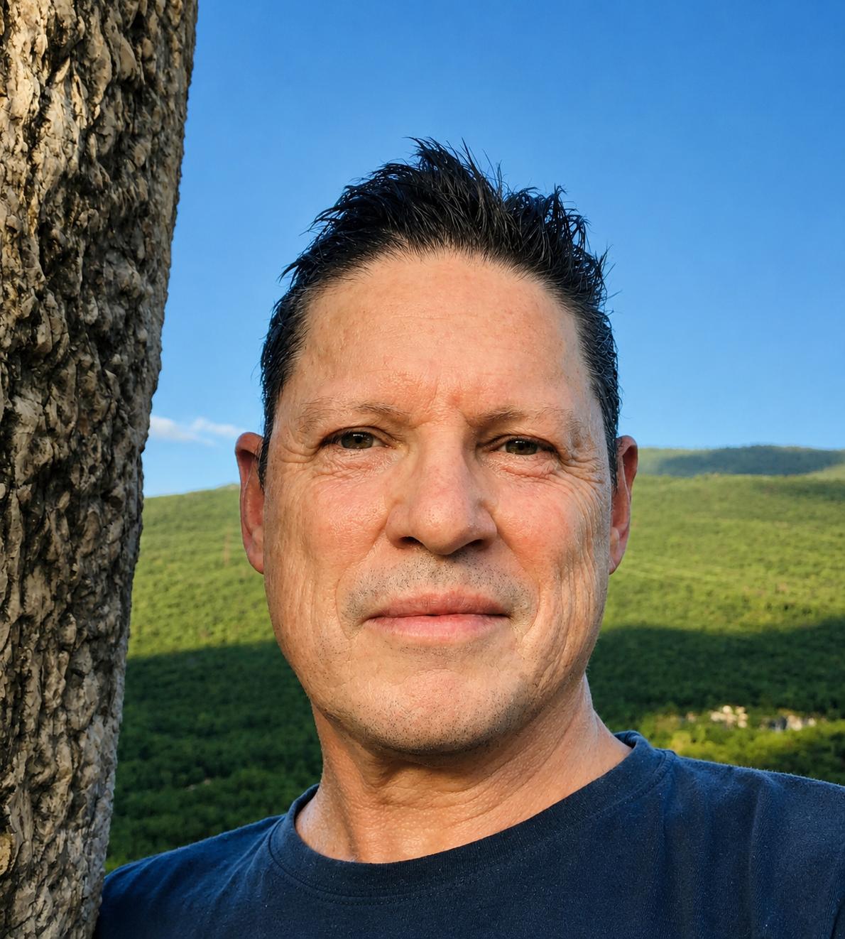 Head and shoulders portrait of a man with short dark hair and a slight smile, standing outdoors beside a tree trunk, wearing a dark T-shirt, with green hills and a clear blue sky in the background.