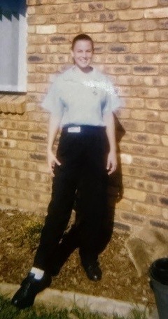 A photograph, slightly aged and with the warm tones characteristic of late 1990s film photography, shows a young woman standing outside a single-storey brick building with a terracotta tiled roof. She is smiling at the camera and wearing a light blue short-sleeved nursing uniform shirt with dark navy trousers and black lace-up shoes, consistent with a student nurse uniform of the era. A name badge is visible on her shirt. She is standing on a brick garden border in front of a garden bed, with two small plant pots visible to her right. The building behind her features large windows with white curtains, suggesting a residential or on-campus accommodation setting.
