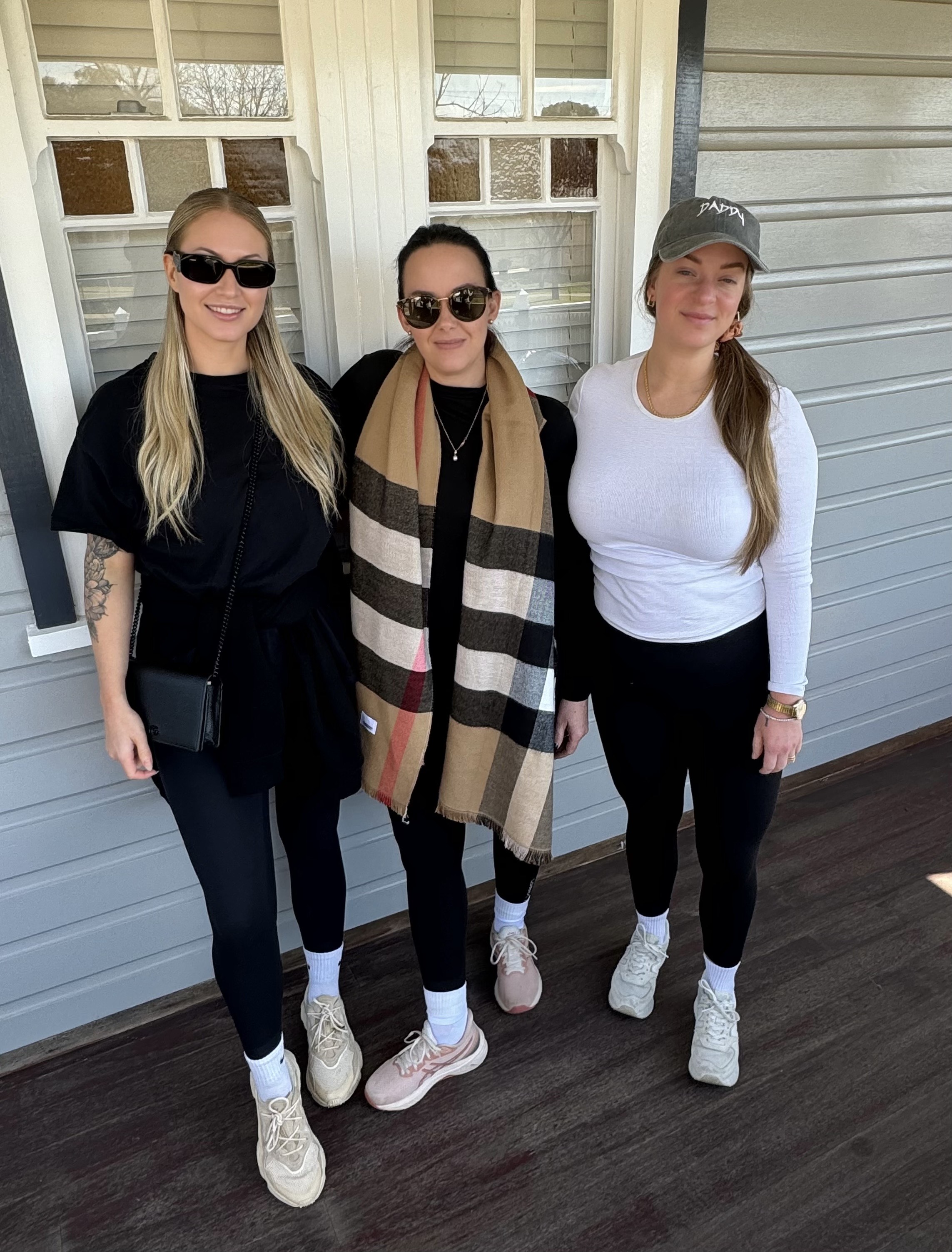 Three women in casual activewear stand side by side on a wooden verandah, smiling at the camera in front of a weatherboard house.