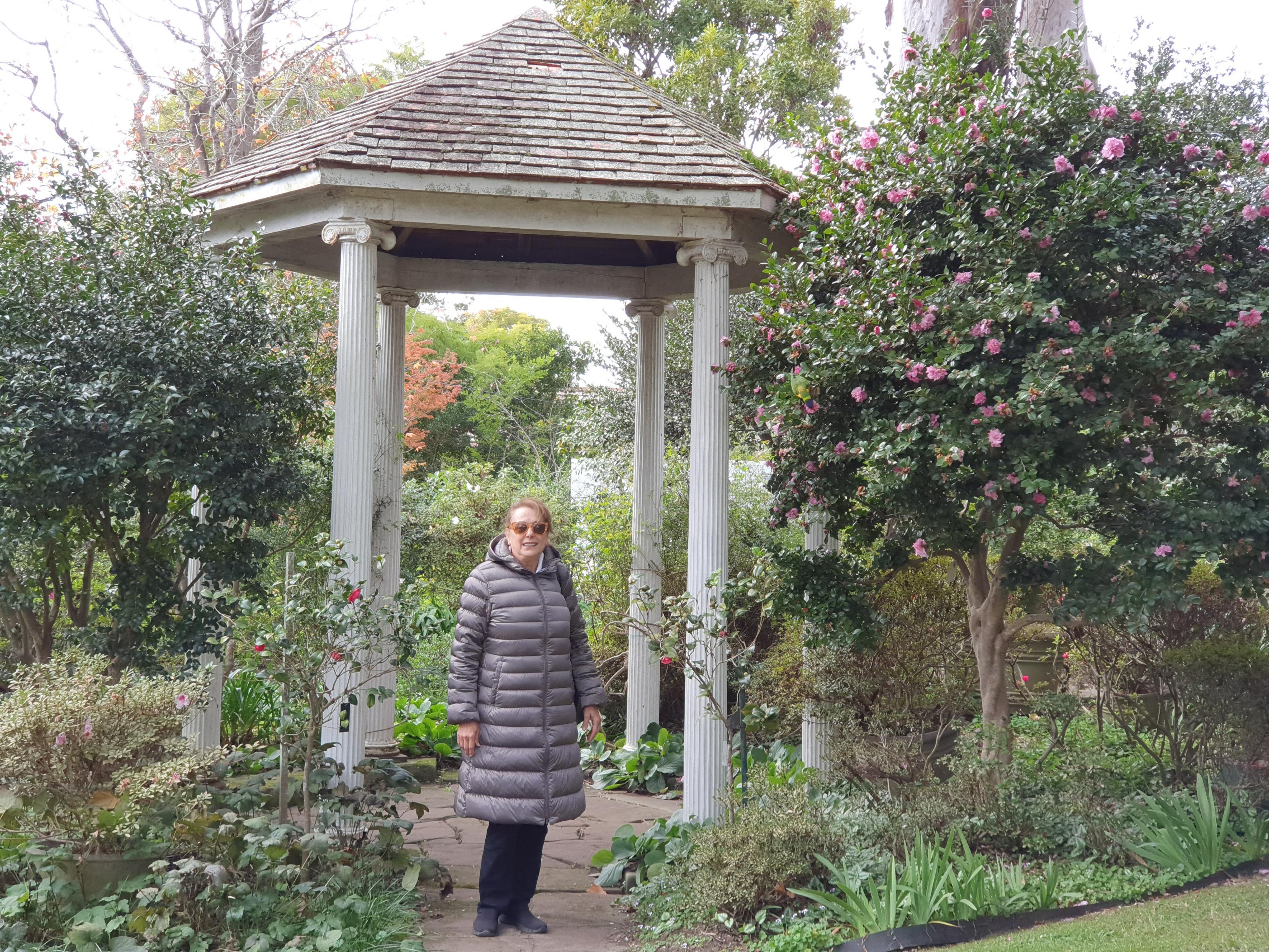 An older woman wearing a long, puffy grey winter coat and sunglasses stands smiling under a small open-sided garden pavilion with a shingled roof supported by four white, fluted columns