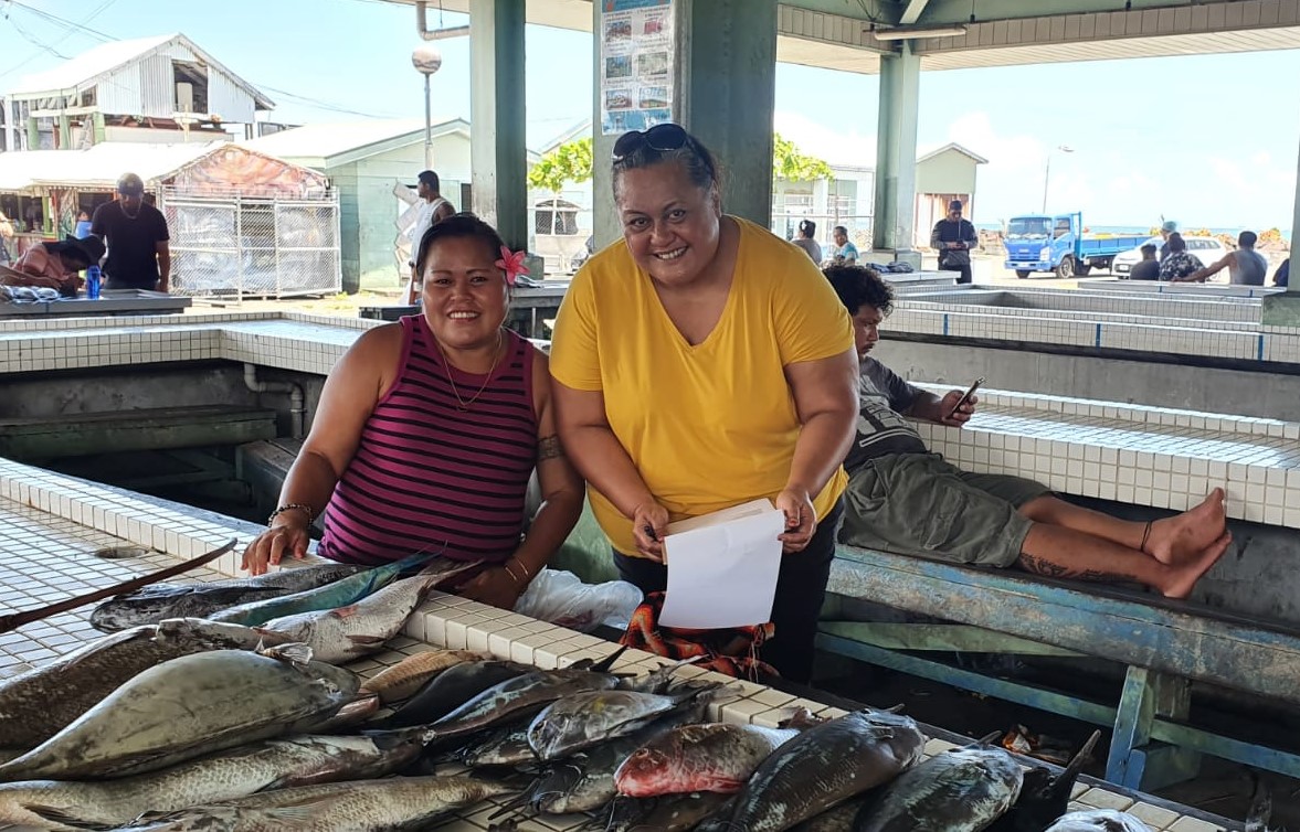 Researcher Dr Fetaomi Tapu-Qiliho smiling with a fish market seller behind a table of colourful market fish. Samoan 