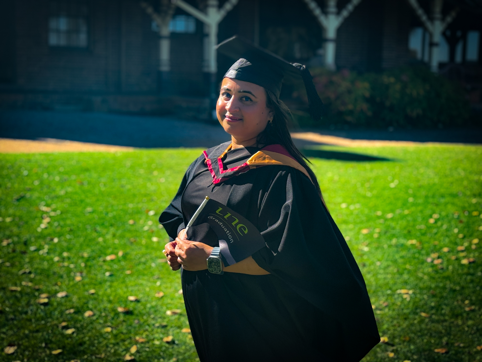 Shila Pokhrel, wearing a black graduation gown with red and gold trim and a mortarboard, stands on the lawn in front of a historic brick building at the University of New England. She holds a small UNE “Graduation” flag in both hands, looking slightly over her shoulder toward the camera with a proud, reflective smile, sunlight highlighting the green grass and autumn leaves around her.