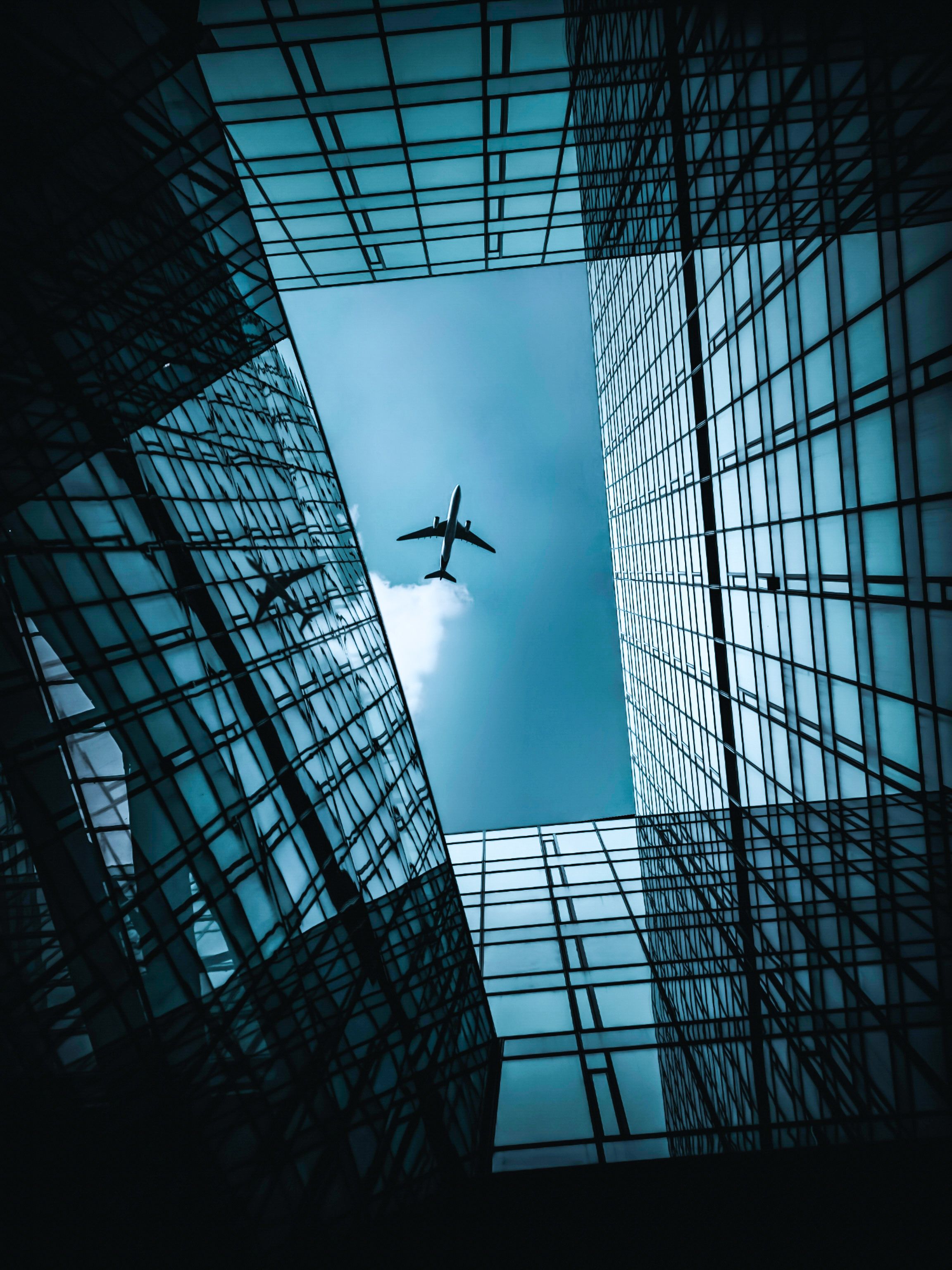 A plane flies over skyscrapers, viewed looking up from the base of the buildings.