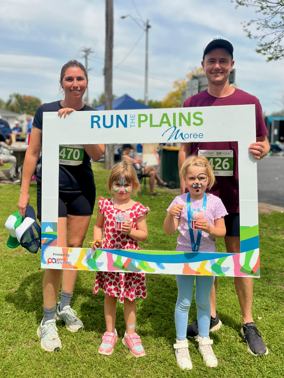 Dr Maelle Morgan and her family pose on a grassy oval at the Run the Plains Moree event, holding a large “RUN THE PLAINS Moree” photo-frame sign while two children with face paint stand in the middle holding icy treats; race bibs are visible on the adults.