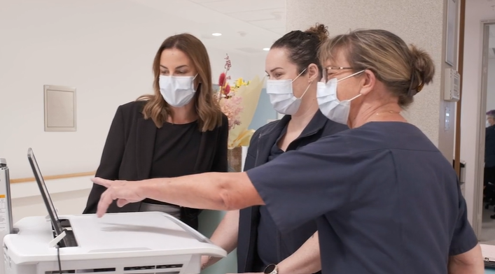 Three women stand together in a bright hospital corridor, gathered around a medical workstation on wheels with a mounted screen. All three are wearing white face masks. The woman on the left is Olivia Tierney, dressed in a black blazer over a black top, with long dark brown hair. She is observing attentively as the woman on the far right, who has greying blonde hair pulled back and is wearing a dark navy clinical uniform, points at the screen on the workstation. A third woman with dark hair, also wearing a dark navy clinical uniform, stands between them. In the background, a bright hospital corridor is visible with a floral arrangement and additional staff members.