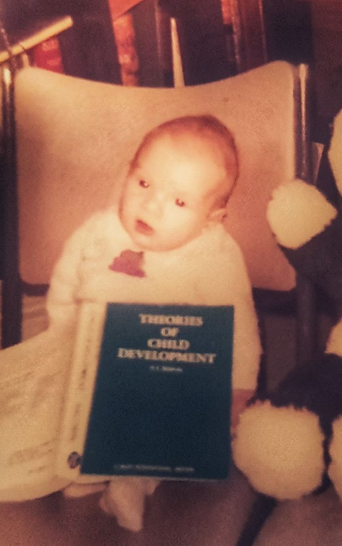 A baby sits propped up on a chair with a book in his lap. The title is 'Theories of Child Development'. The child is smaller than the teddy bear next to him.