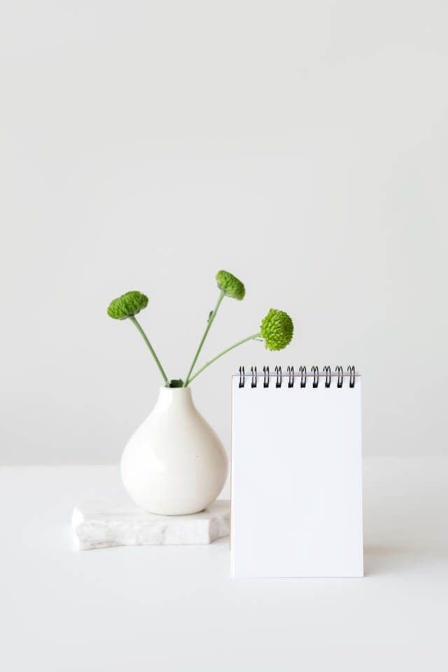 White vase with green flowers on a white desk with a white notebook bound by a black spiral