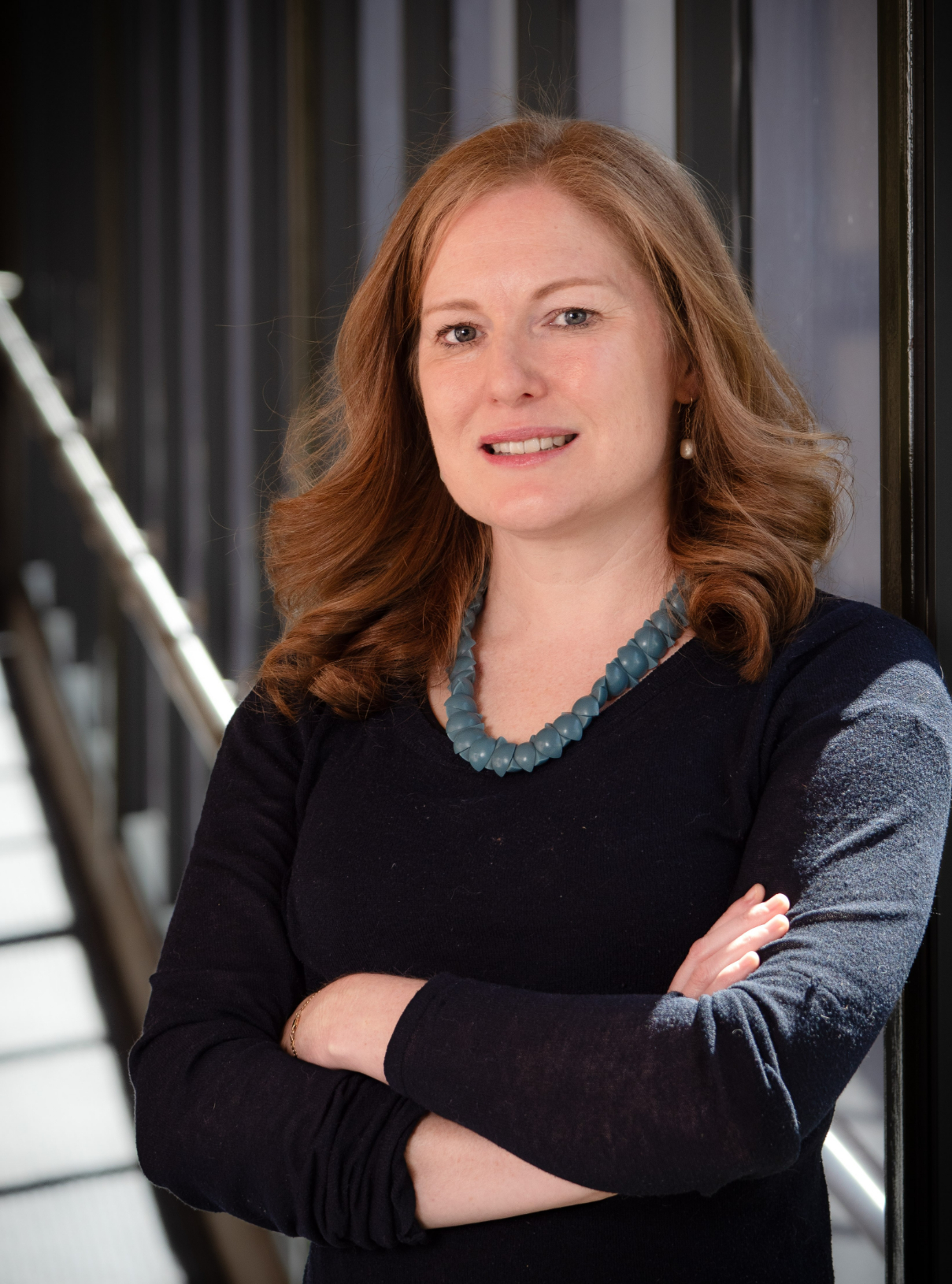 Red-haired woman standing in a glass-lined corridor with arms crossed, wearing a dark long-sleeve top, teal chunky bead necklace, and small drop earrings, looking at the camera with a slight smile.