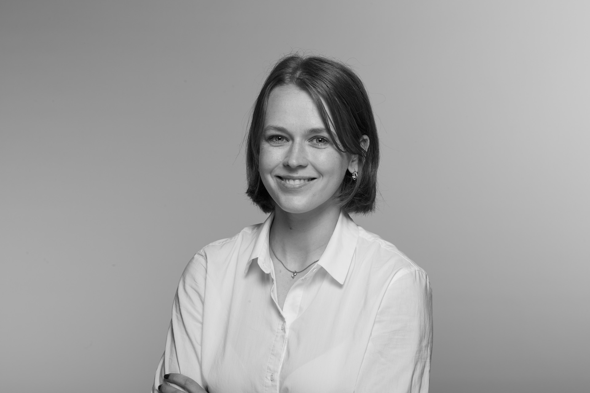 A black-and-white studio portrait of a young woman with a neat, chin-length bob haircut, smiling softly at the camera. She is wearing a light-coloured button-up shirt with the sleeves rolled up and a small necklace, standing against a smooth, plain background with her arms loosely crossed in front of her.