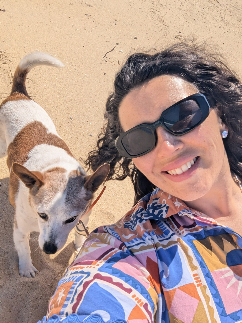 Smiling person wearing dark sunglasses and a colourful patterned shirt takes a close-up selfie on a sunny beach, with a small white and brown dog standing beside them on the sand.