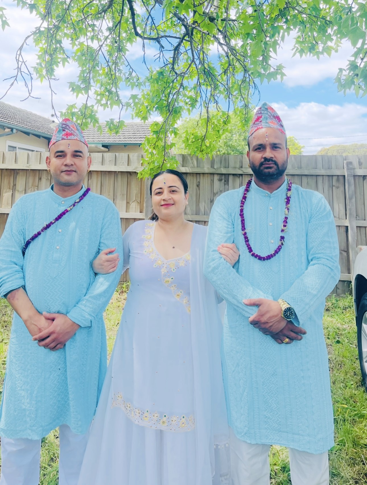 Shila Pokhrel stands outdoors between her two brothers under a leafy green tree, all three dressed in coordinated traditional outfits. Shila wears a flowing white dress with delicate yellow and gold embroidery and a matching shawl, while each brother wears a light blue kurta with white trousers, a traditional patterned topi hat and a purple flower garland. They link arms and face the camera, with a timber fence, lawn and blue sky in the background.