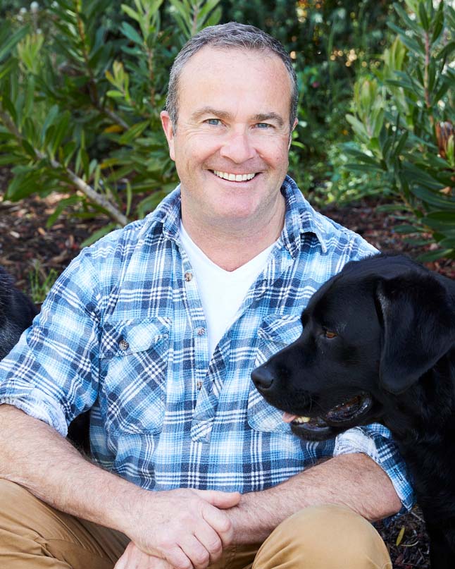Paul McGreevy smiling at the camera. He is wearing a blue, plaid shirt with brown pants, and there is a black labrador beside him.