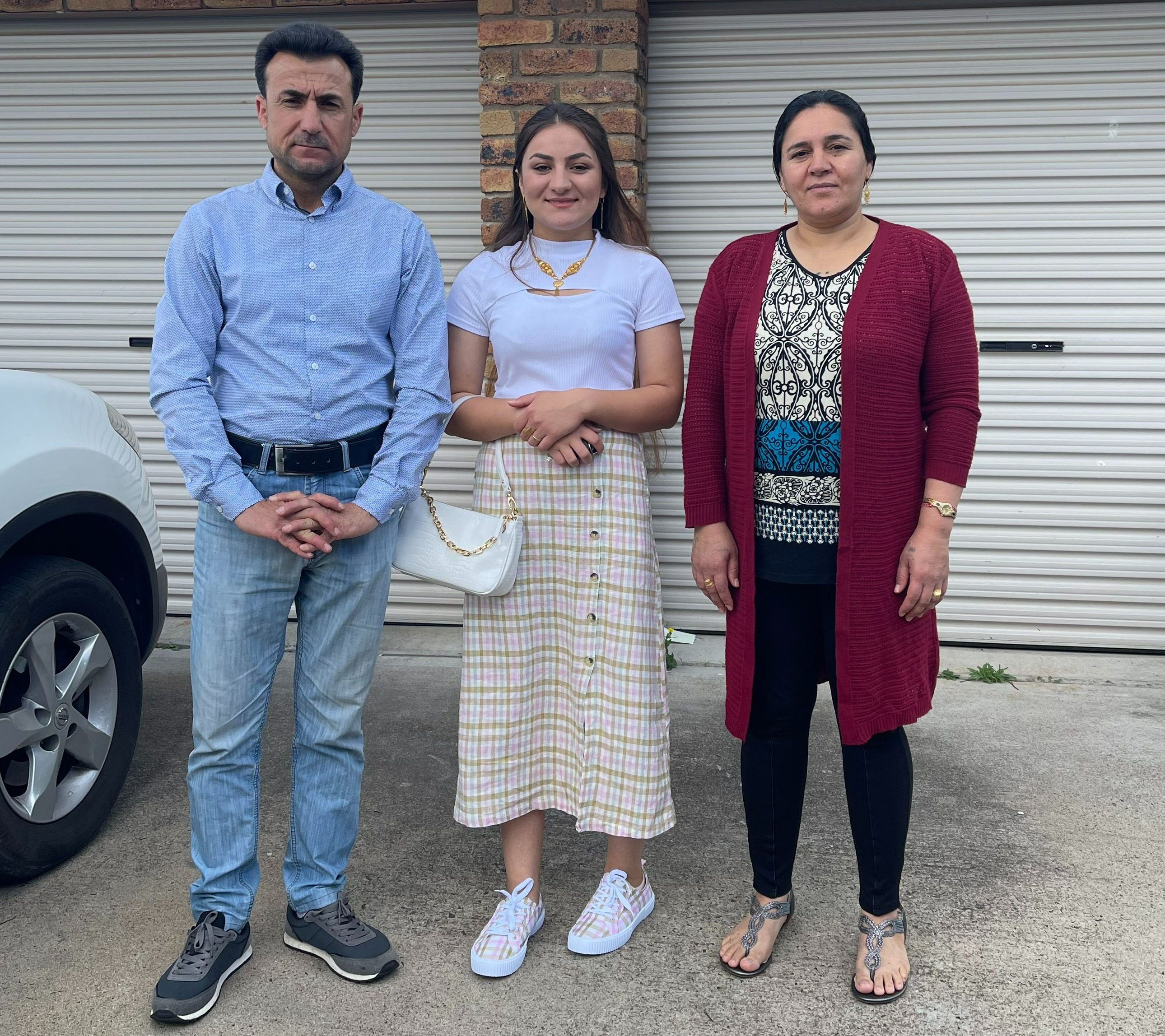 Ezidi family of older Mother and Father with teenage daughter standing in driveway at the front of their house.