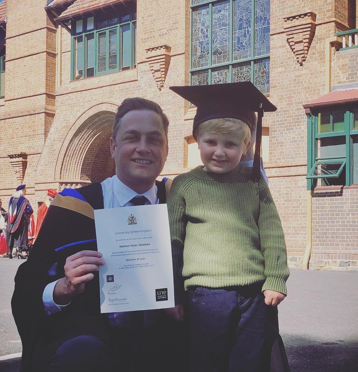 Man in graduation gown holds toddler son who is wearing trencher on head
