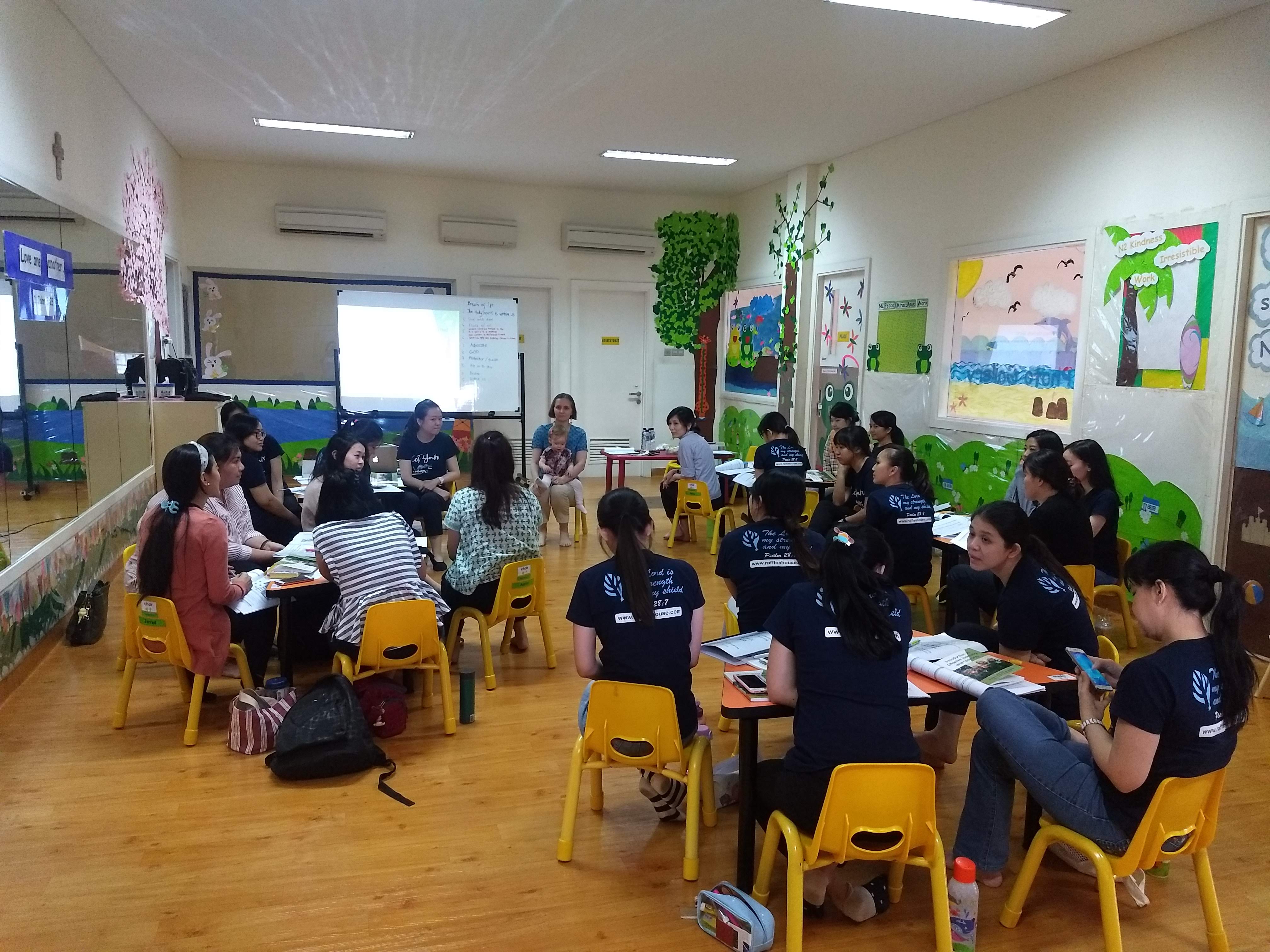 Beth Boland sits at a whiteboard at the front of a classroom with a baby in her lap, talking to a class of adult learners in an early childhood education classroom