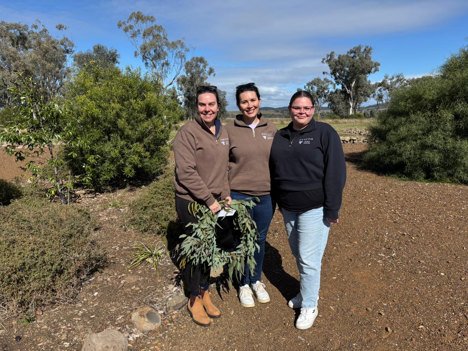 Three women stand together smiling on a sunny day in a bushland garden, wearing NERCHA-branded jumpers. The two on the left wear brown tops and hold eucalyptus branches, while the woman on the right wears a black jumper and light blue jeans. Low shrubs and taller gum trees surround them under a bright blue sky with scattered clouds.