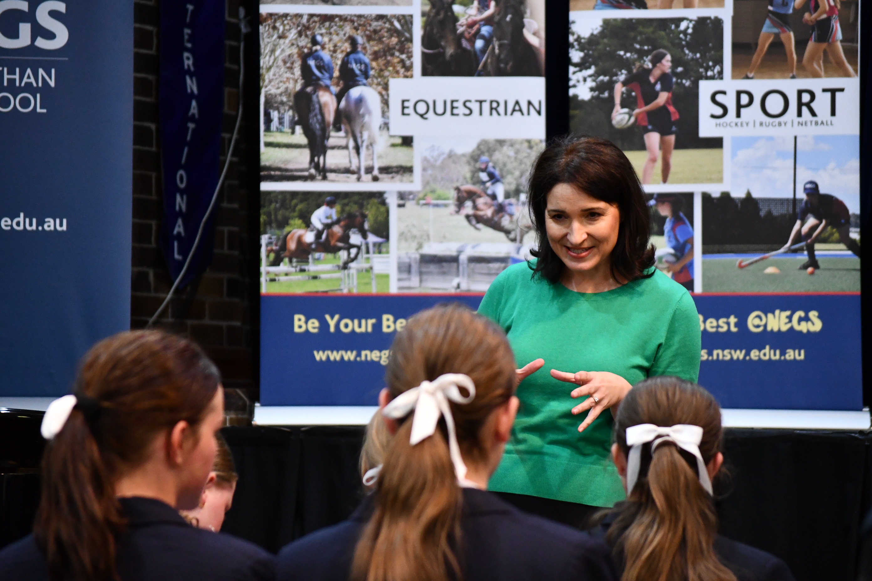 Dr Mary Kaspar stands at the front of a school hall speaking to a small group of teenage girls in uniform. She is wearing a bright green top and gesturing with her hands as the students, their hair tied back with white ribbons, listen closely. Behind her are banners featuring equestrian and sports photos and the slogan “Be Your Best @NEGS.”