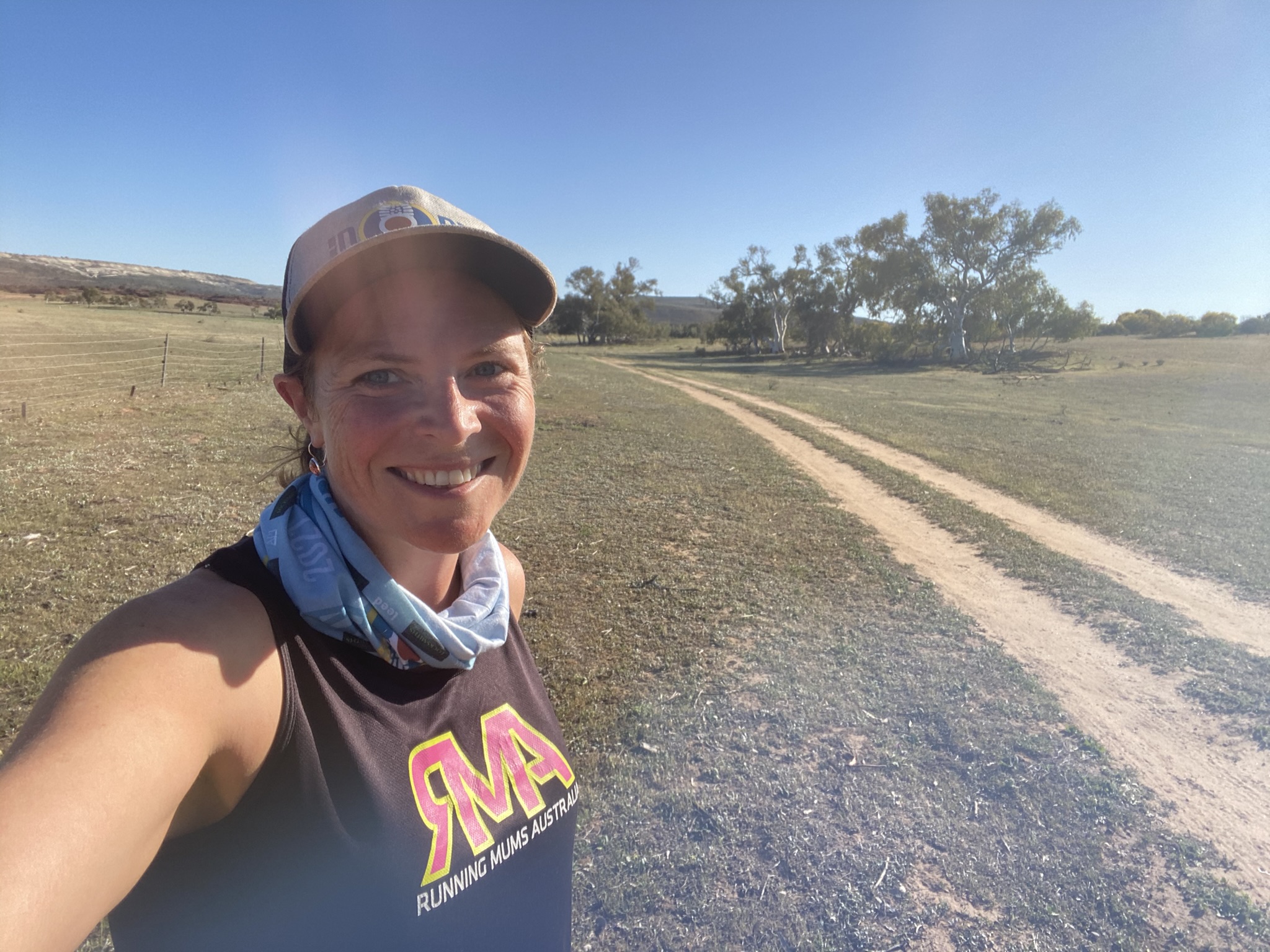 Julie Clark smiles at the camera while standing on a dusty farm track under a clear blue sky, wearing a cap, neck buff and “Running Mums Australia” singlet, with open paddocks and scattered trees in the background.