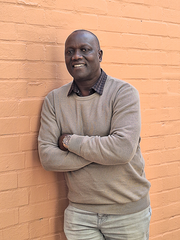 Portrait image of Sam Onapa leaning against an orange brick wall. 