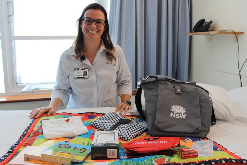 Olivia Tierney, a woman with long dark brown hair and glasses, smiles warmly at the camera while standing behind a hospital bed covered in a colourful patchwork quilt. She is wearing a light blue striped shirt and a NSW Health staff lanyard and ID badge. Spread across the bed in front of her are a range of maternal and newborn care items, including a red first aid kit labelled 