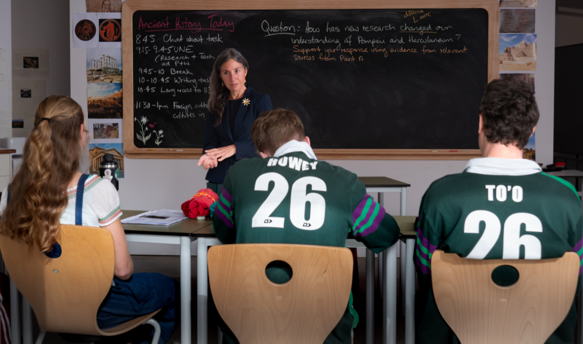 Woman standing before a blackboard in a classroom talking to three students with their backs to the camera.