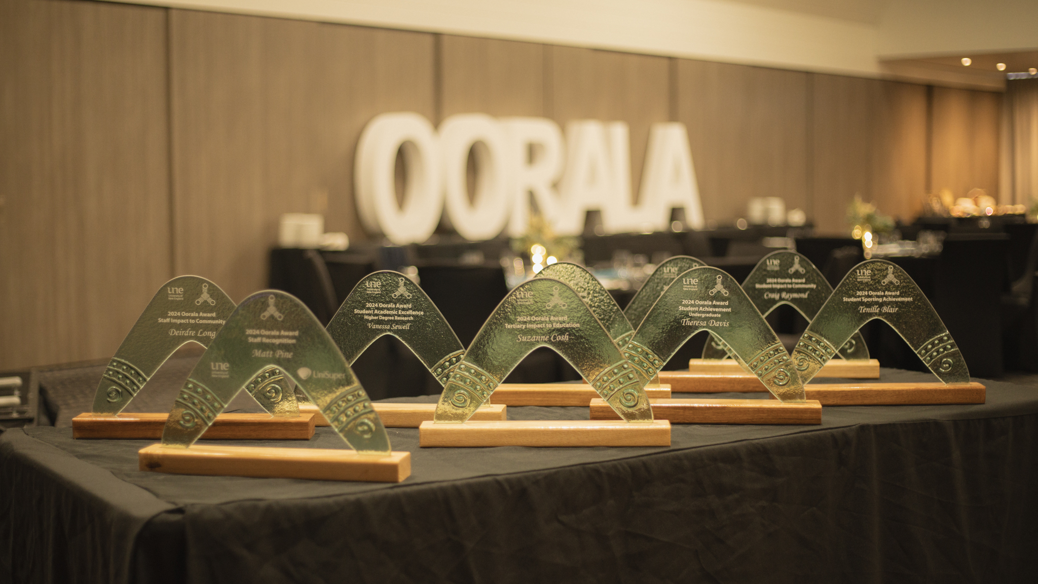A row of glass boomerang-shaped awards with wooden bases displayed on a black tablecloth at an awards ceremony. Each award is engraved with the recipient’s name and category. In the background, large white foam letters spell ‘OORALA’ in front of a dimly lit dining area.
