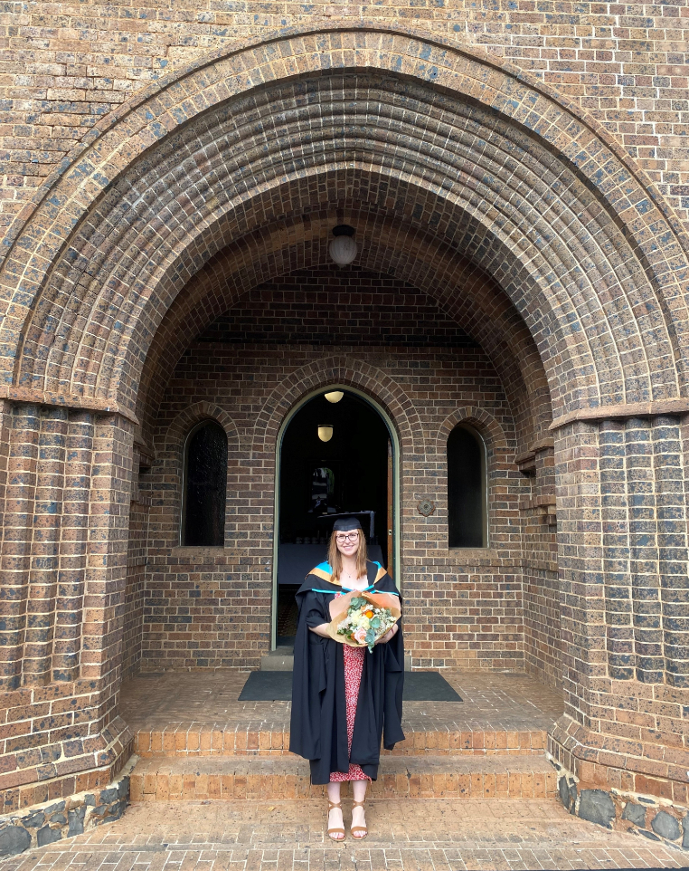 Brianna Carney standing in an archway with a bunch of flowers at her UNE graduation.