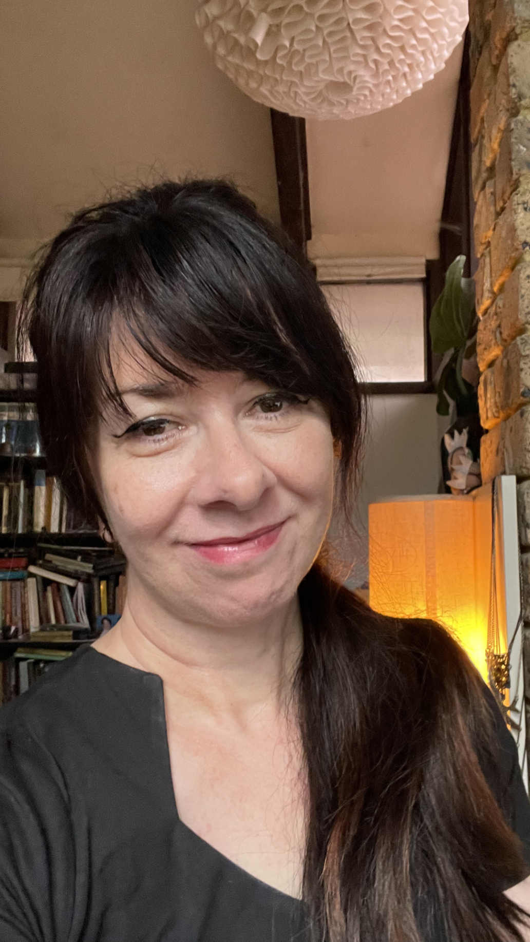 Portrait of Danielle Akayan smiling at the camera in a warmly lit room, with bookshelves, an exposed brick wall and a glowing lamp in the background.