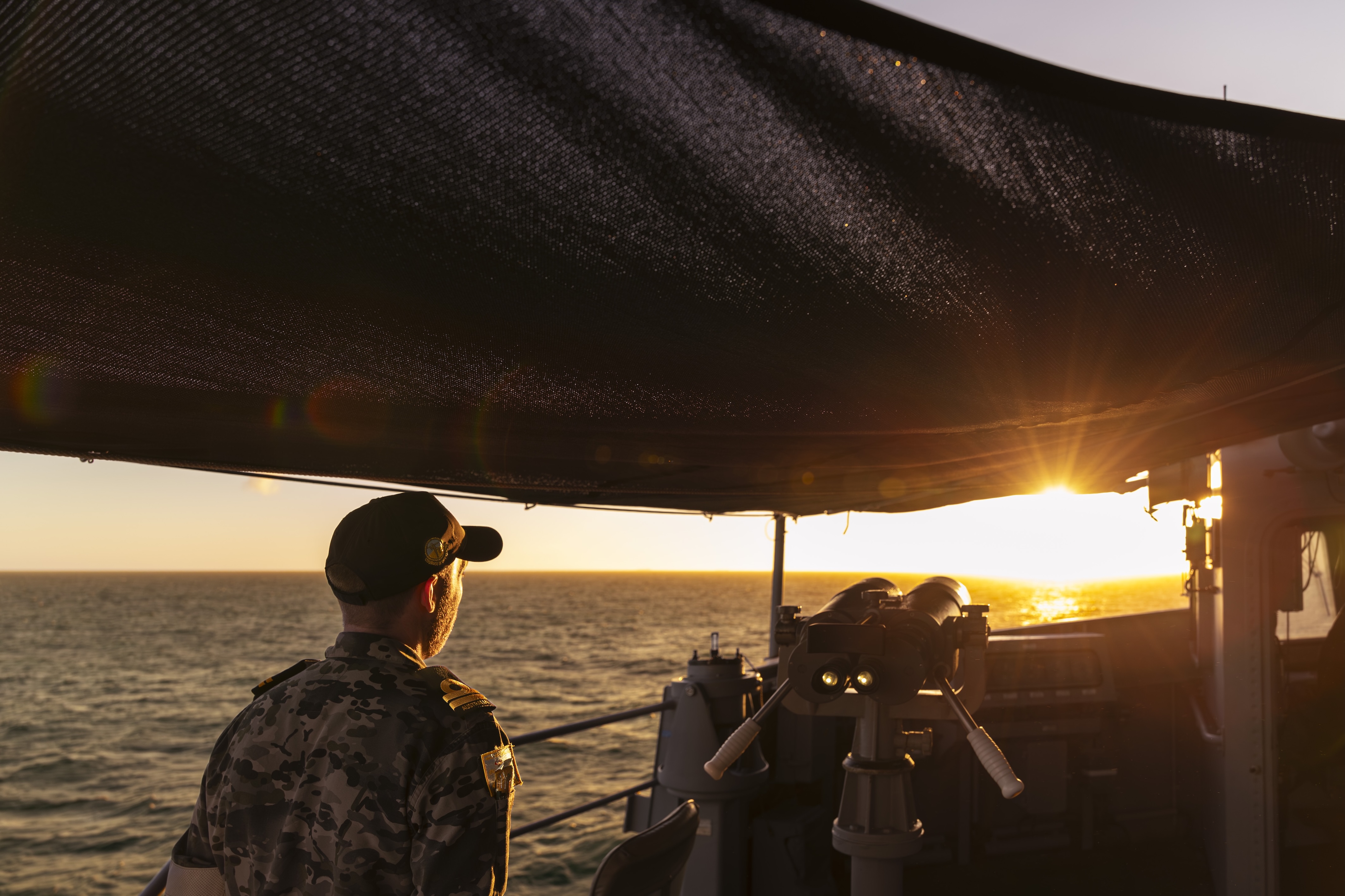 And Australian Navy member on a ship looking out at a sunset over the water.
