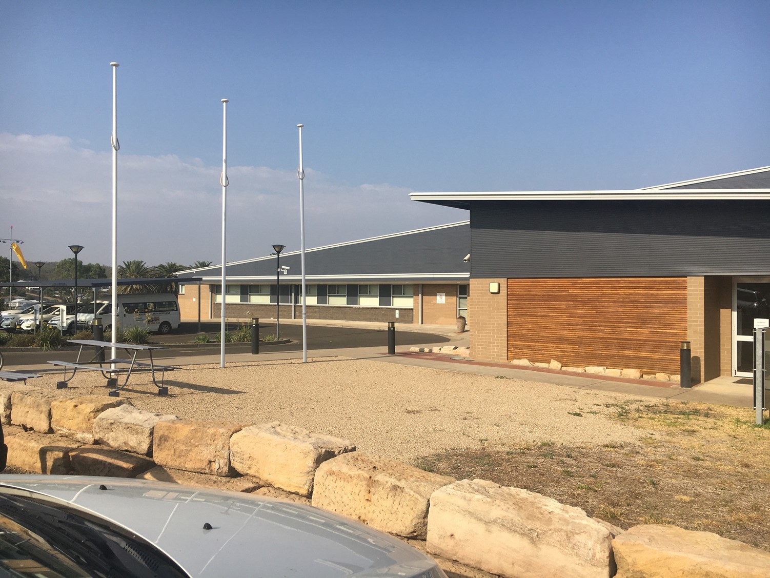 Exterior view of a modern, single-storey regional facility featuring a combination of brick, timber cladding, and dark grey metal roofing. Three tall, empty flagpoles stand in a gravel forecourt bordered by large sandstone rocks. A metal picnic table sits in the foreground. Several vehicles, including a white van, are visible in a car park to the left, shaded by a covered structure. Palm trees and low shrubs are visible in the background under a clear blue sky with light haze on the horizon.