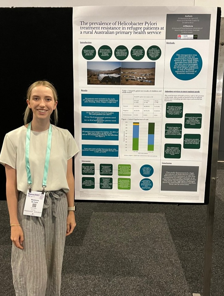 Madeleine de Carle, a young woman with blonde hair wearing a white short-sleeved top, grey striped wide-leg pants, and a teal conference lanyard, stands smiling beside her academic research poster at the WONCA 2023 conference. The poster is titled 