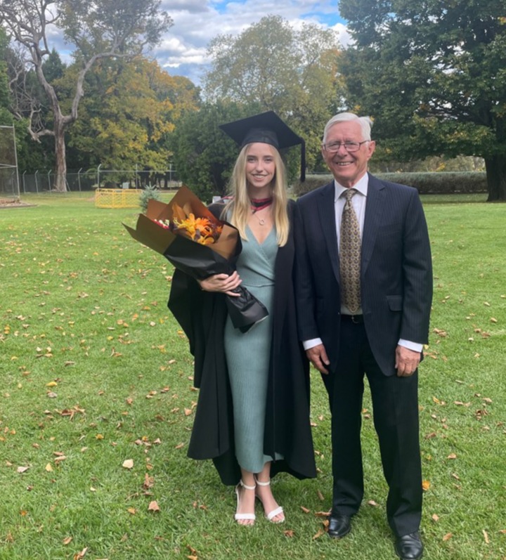 Madeleine de Carle, a young woman with long blonde hair wearing a black academic graduation gown and mortarboard cap over a sage green midi dress and white heeled sandals, stands smiling on a grassy lawn beside her grandfather, Dr David de Carle, an older man in a dark pinstripe suit and patterned tie with glasses. Madeleine holds a bouquet of sunflowers wrapped in dark paper. Autumn-coloured trees and a partly cloudy blue sky are visible in the background, with fallen leaves scattered across the lawn.