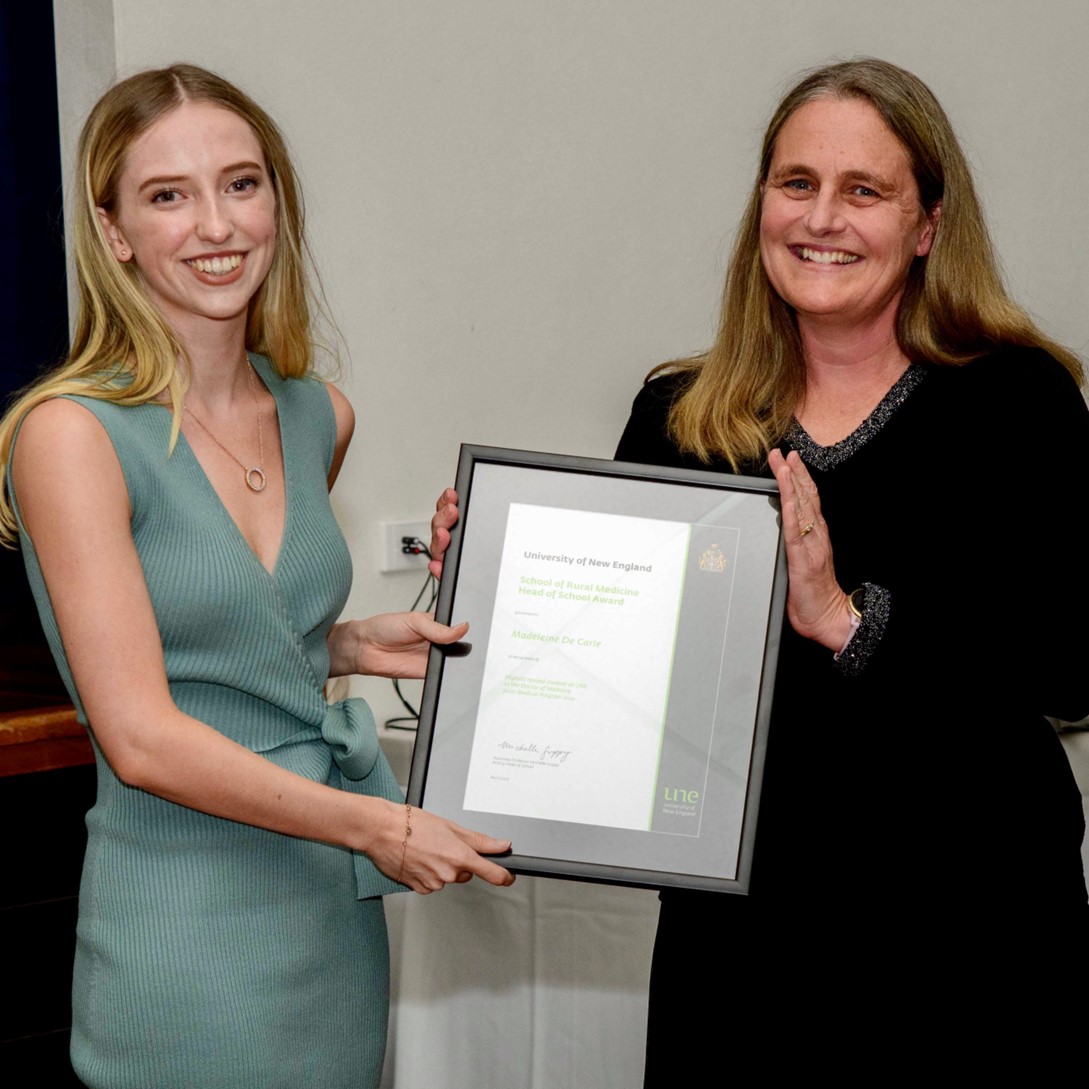Madeleine de Carle, a young woman with long blonde hair wearing a sage green sleeveless ribbed dress and a delicate gold circle necklace, smiles as she holds a framed certificate alongside her mentor and co-author, a woman with long brown hair wearing a black top with subtle sparkle detail. The framed certificate reads 