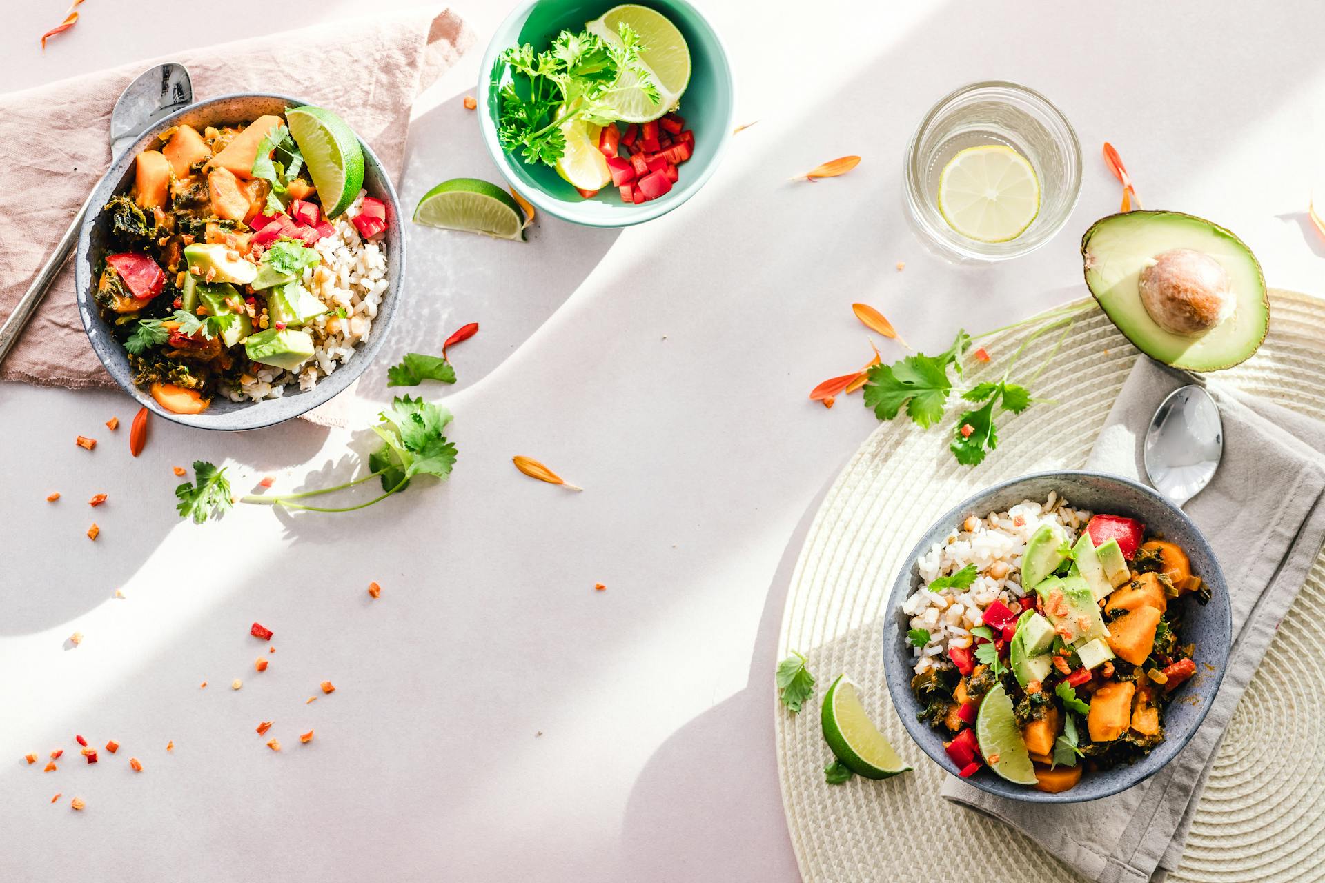 Overhead view of two colorful grain bowls with roasted sweet potato, red capsicum, fresh cilantro, lime wedges, and avocado, styled on a white surface with fresh herbs and lemon water, demonstrating demonstrating nutrient-rich, colourful eating as discussed by Dr Vandana Gulati.