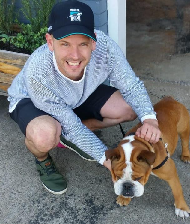 Person crouching on a footpath, smiling at the camera while holding and gently patting a brown and white bulldog on a leash, next to a wooden planter box and a building entrance.