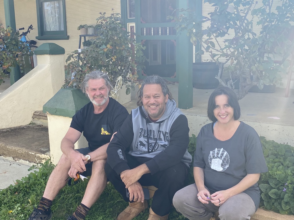 Three adults sit together on a low step in front of a weathered house verandah, smiling at the camera. On the left, a man with grey hair and beard in a black T‑shirt and patterned socks rests his hands on his knees. In the middle, a man with shoulder‑length hair and stubble wears a grey and black “Bulldogs” hoodie. On the right, a woman with dark bobbed hair wears a loose grey T‑shirt with a circular graphic and light pants, hands clasped in her lap. Behind them are green-painted posts, potted plants, and leafy shrubs framing the porch.