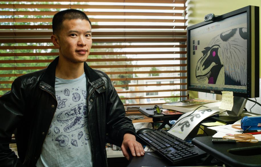 Tommy Leung stands at his computer, which shows one of his parasite illustrations in black and white with purple highlights. He wears a black leather jacket over a T-shirt with drawings of fossils on it.