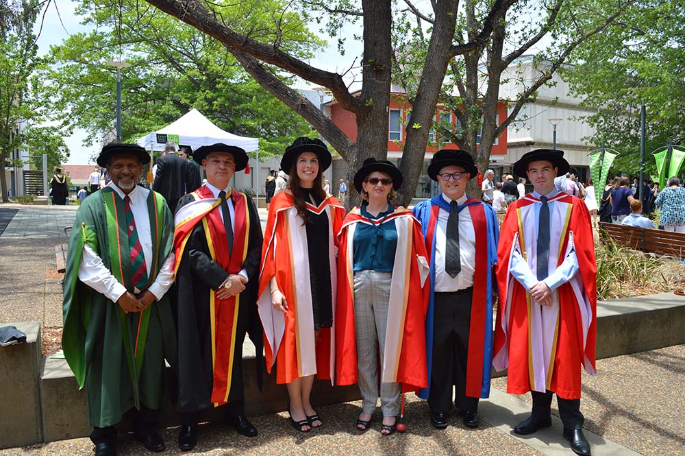 Image of Sinead Henderson standing in a group with Linda Agnew at her graduation ceremony. They are wearing red robes, and there is a large tree in the background.