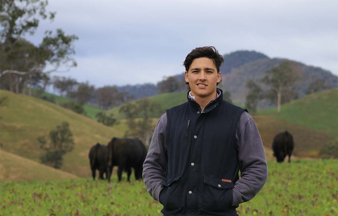 Image shows Angus Dingley smiling at the camera. Angus cattle are in a green paddock in the background. 