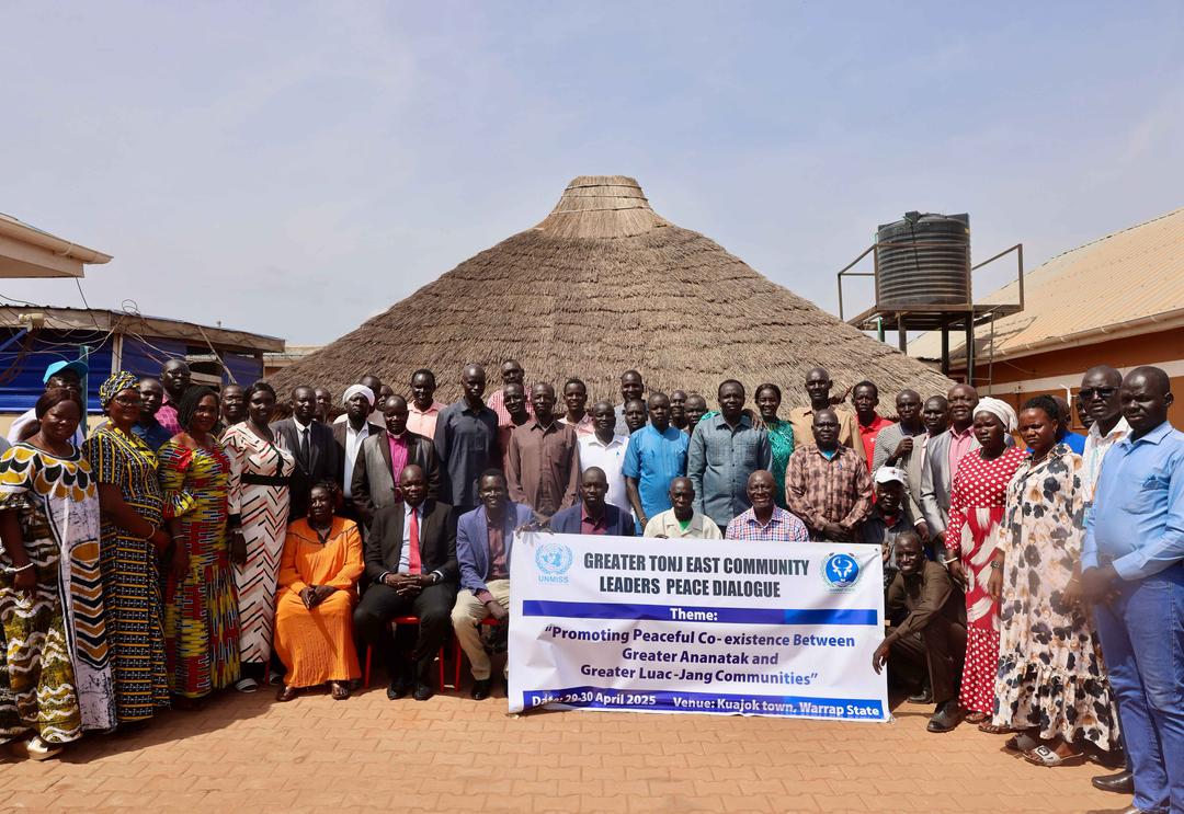 Large group of community leaders and participants gathered outdoors in front of traditional thatched hut, holding banner for Greater Tonj East Community Leaders Peace Dialogue in Kuajok town, Warrap State