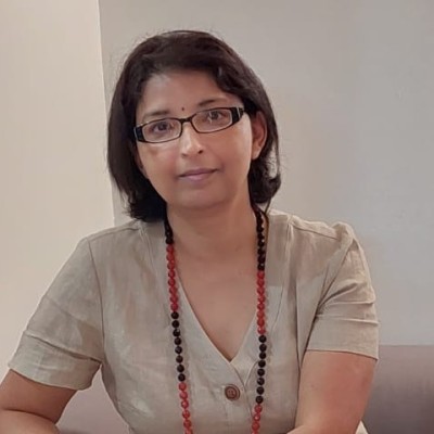 A person with short dark hair and glasses sits indoors against a light-coloured wall, looking calmly toward the camera. They are wearing a beige short-sleeved top and a long beaded necklace with alternating dark and reddish beads, with their hands resting in front of them.
