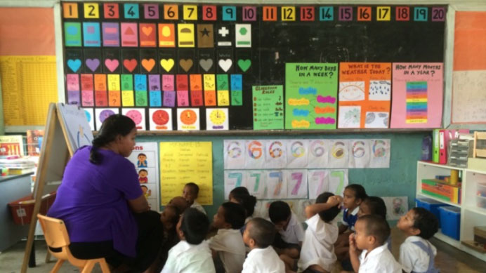 A colourful Nauru classroom, young students sit on the classroom floor and listen to their teacher.