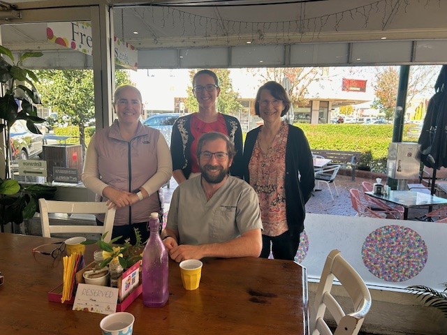 Three female doctors stand behind male medical student sitting at a table in a cafe.