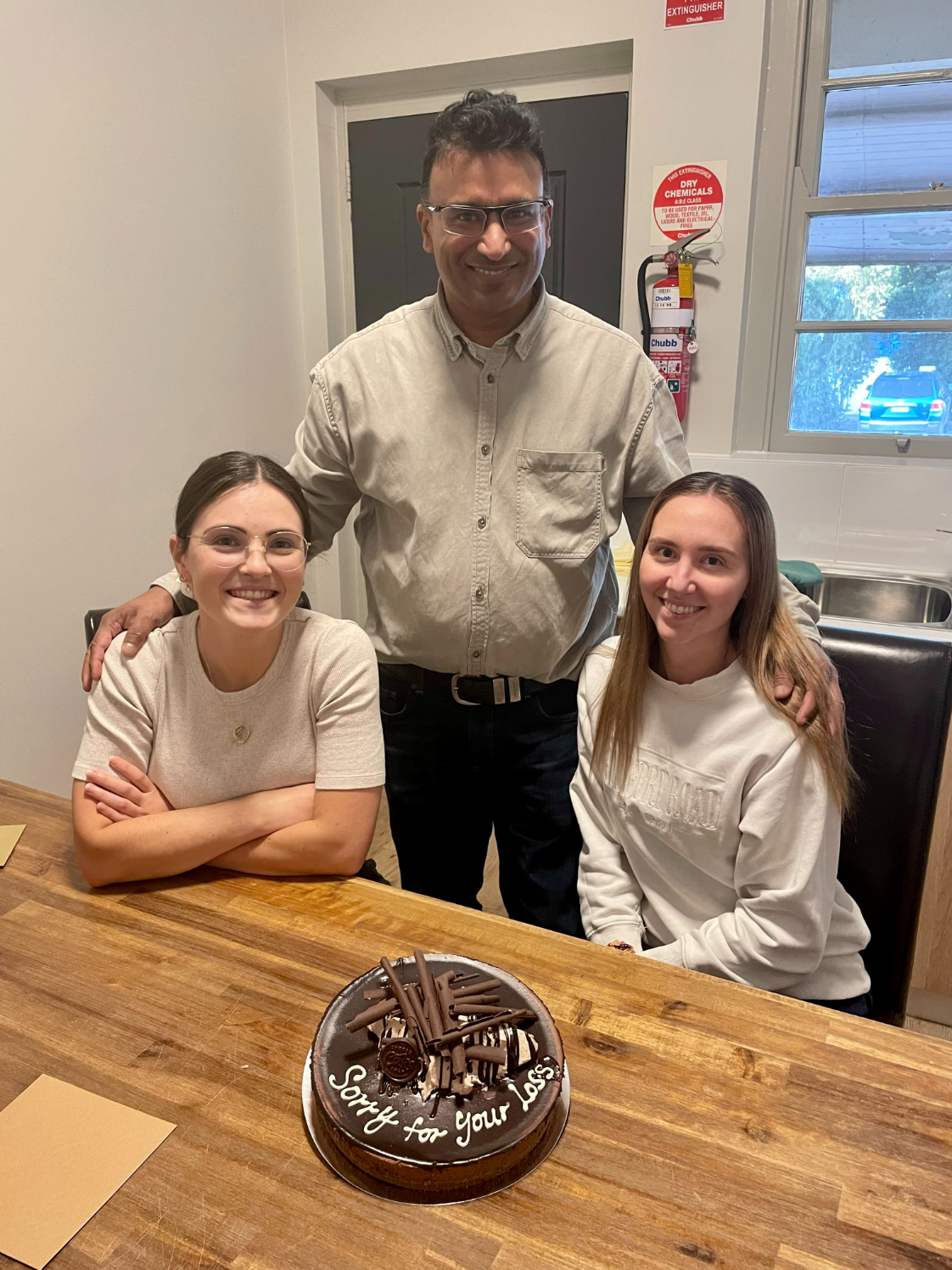 Two young female med students sit either side of a man at a dining table and pose for a photo in front of a birthday cake