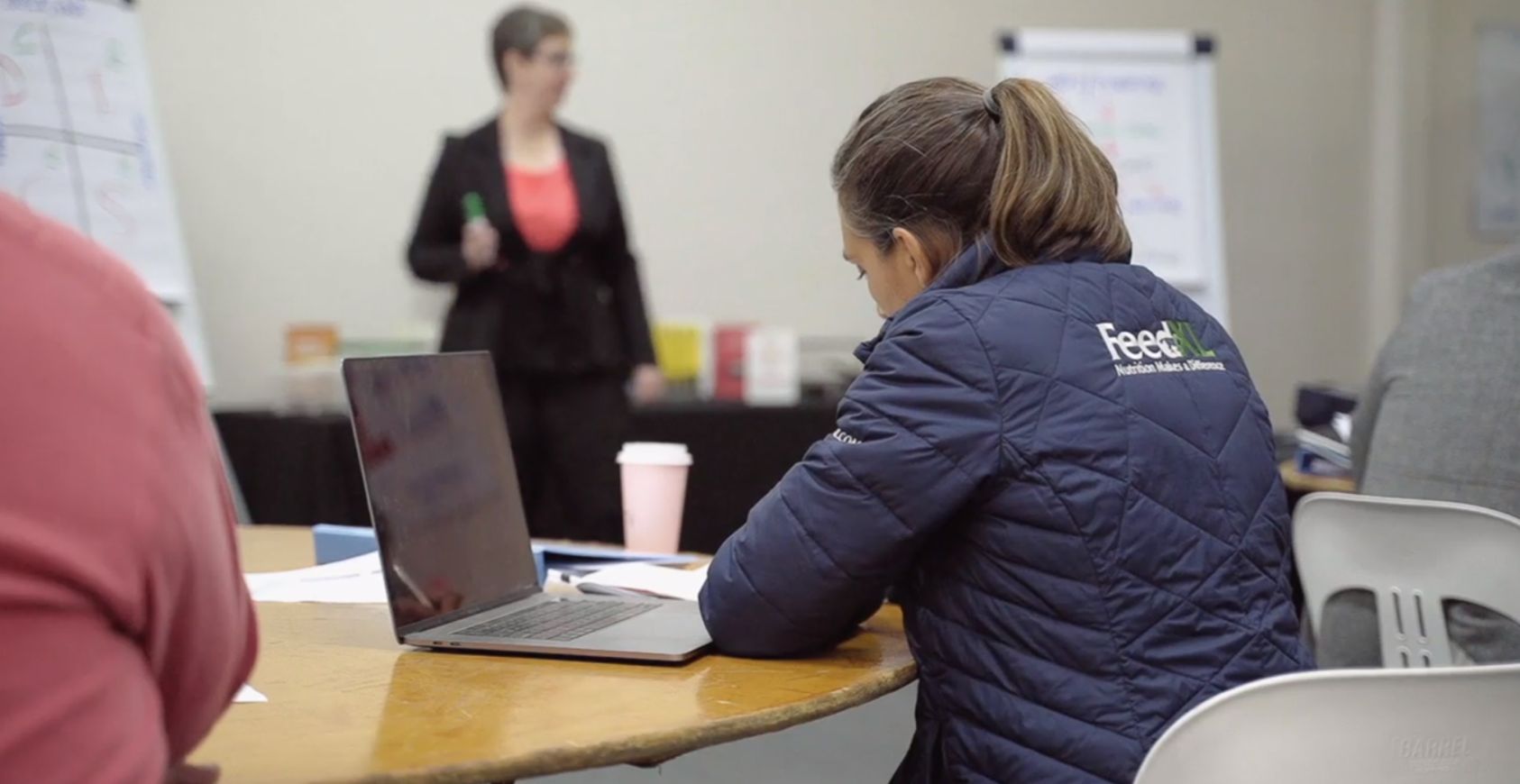 A woman sits at a laptop, her back to the camera, as a female presenter addresses the group.