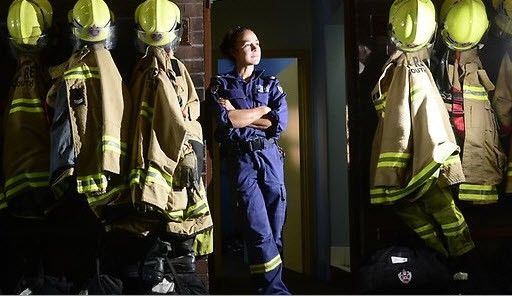Tara Lal stands in firefighting uniform, in the doorway of a brick wall. Her arms are crossed and she's looking off to her left. On the walls hang a row of firefighting uniforms.