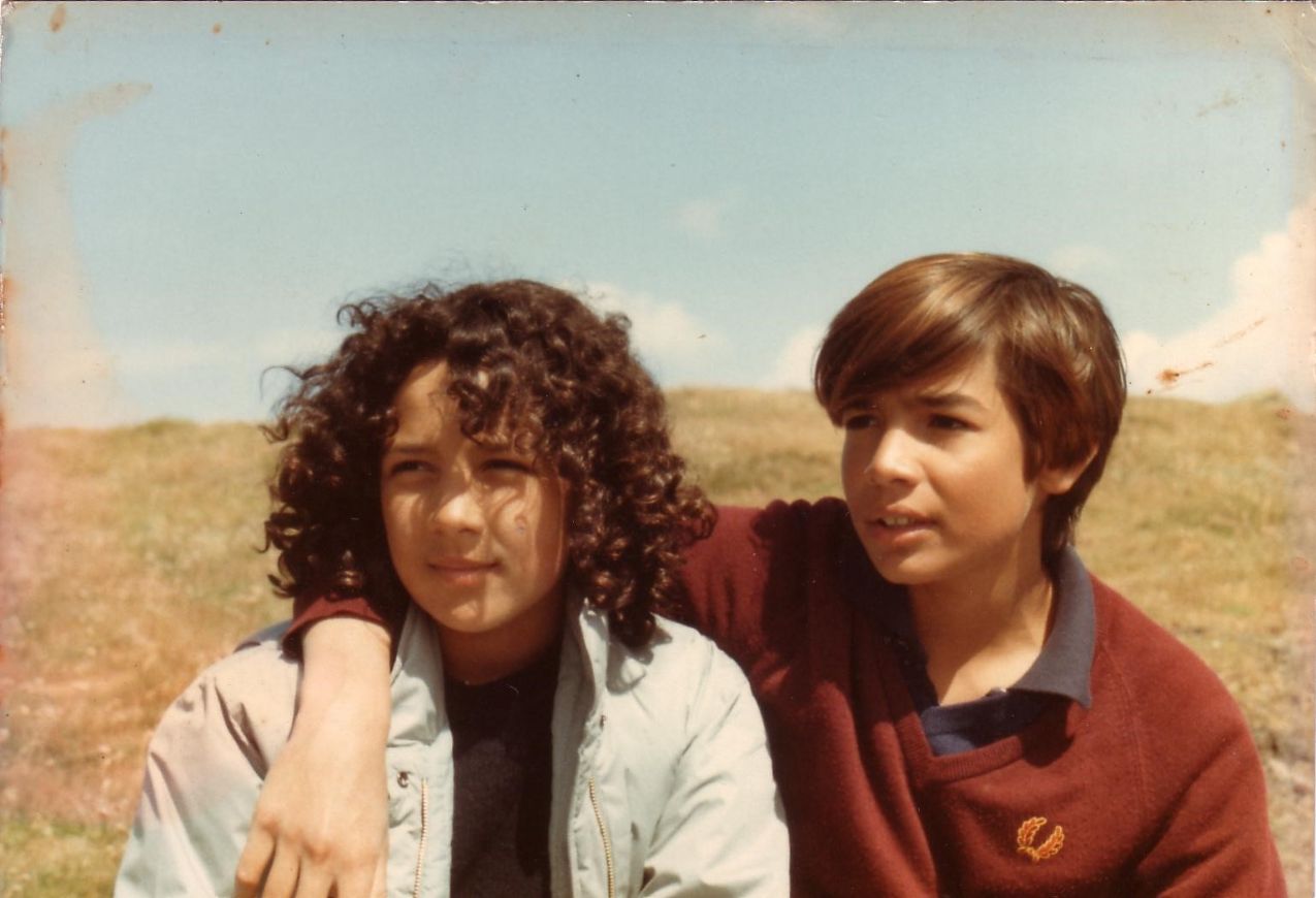 A young Tara Lal and her brother Adam. Photo is in colour with sepia tones, and shows a girl with dark, curly hair and a boy with shiny chestnut hair. They are standing outdoors in an open field and looking off to their right. His arm is around her shoulders.