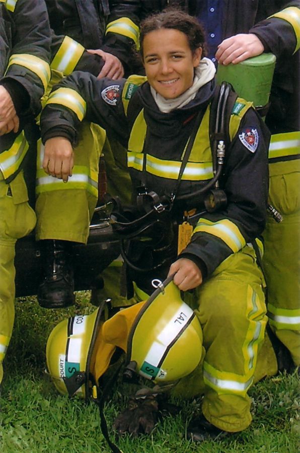 Tara Lal kneels among colleagues, wearing her black and yellow firefighting uniform. She holds a yellow helmet and is smiling at the camera.